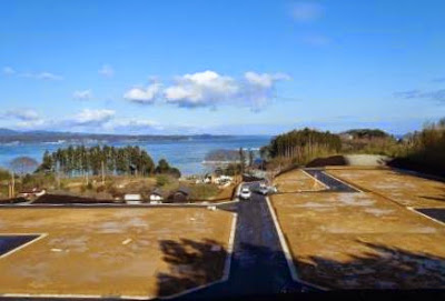 整備された区画と舗装道路の向こうに海が広がり、周囲には林と住宅地が見える高台の造成地からの展望を写した写真