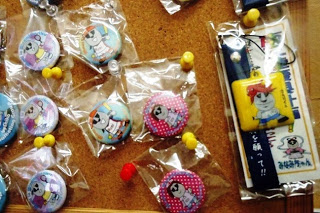 透明な袋に入った色とりどりのモアイのキャラクター缶バッジとストラップがコルクボードに掲示されている写真