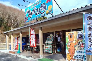 「南三陸直売所 みなさん館」と書かれたカラフルな看板がある建物の前に、のぼり旗や販売物が並び顔出しパネルが置かれている直売所の外観写真