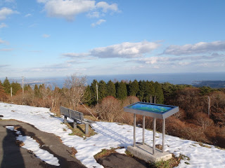 雪が積もった田束山の展望台から、ベンチと案内板越しに冬枯れの木々とその先に海が広がる写真