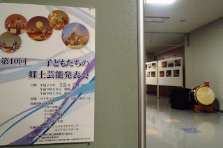 壁に「第10回 子どもたちの郷土芸能発表会」と書かれた案内ポスターが貼られており、奥の廊下には展示された写真や和太鼓が置かれている様子を写した写真