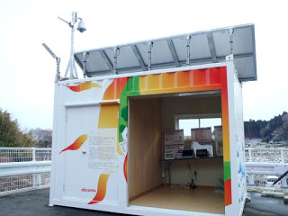 赤、オレンジ緑などで太陽のような絵が描かれた白いコンテナが屋外に設置されており、解放されたドアの中には複数の機器が見え、屋根の上にはソーラーパネルが取り付けられている写真