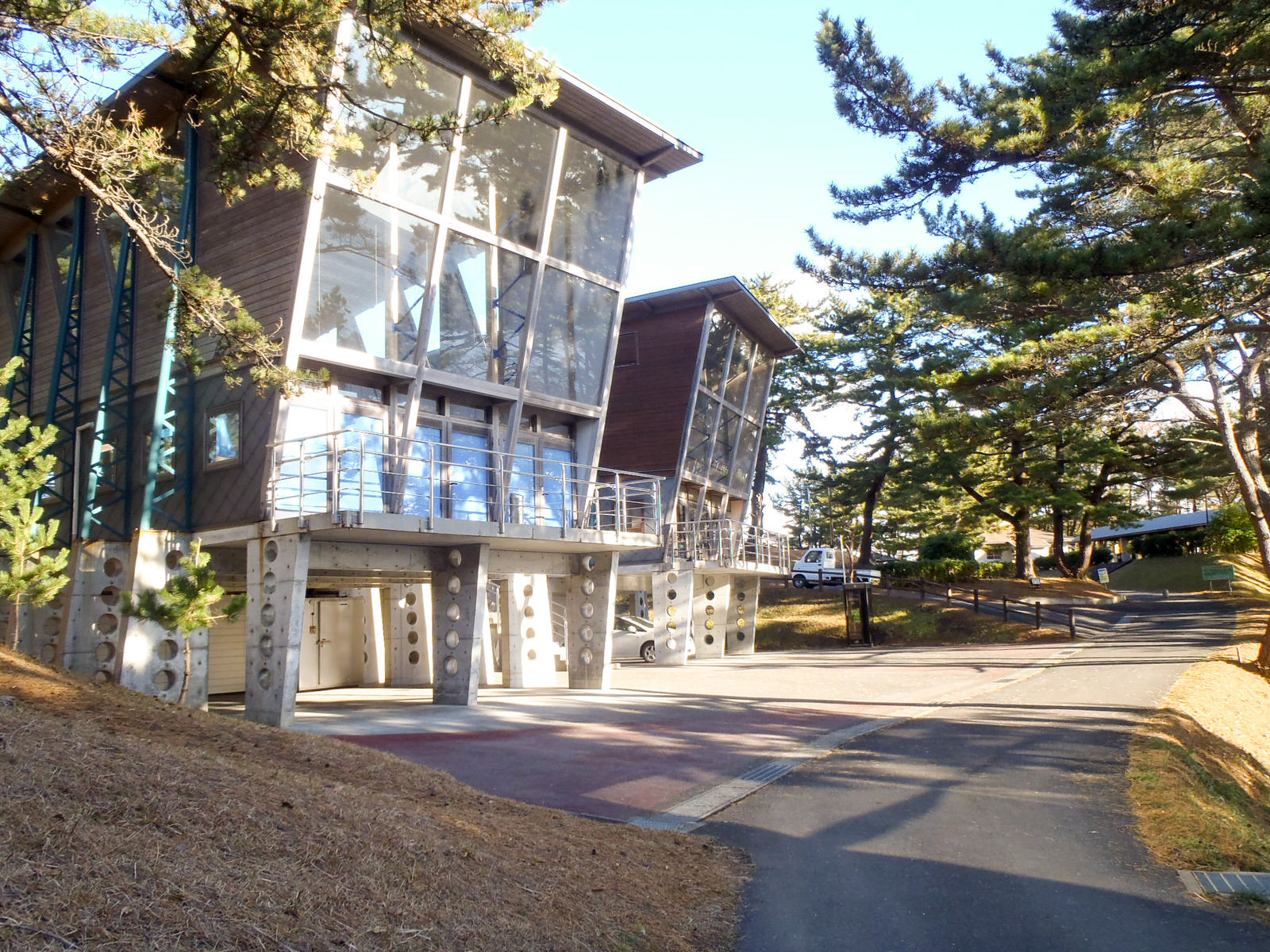 丸い穴のあいた柱に支えられた、大きなガラス窓と斜めの屋根が特徴的なモダンな高床式の建物が2棟並んで建ち、周囲を松の木々に囲まれた中で、舗装された道がゆるやかに続いている施設の写真