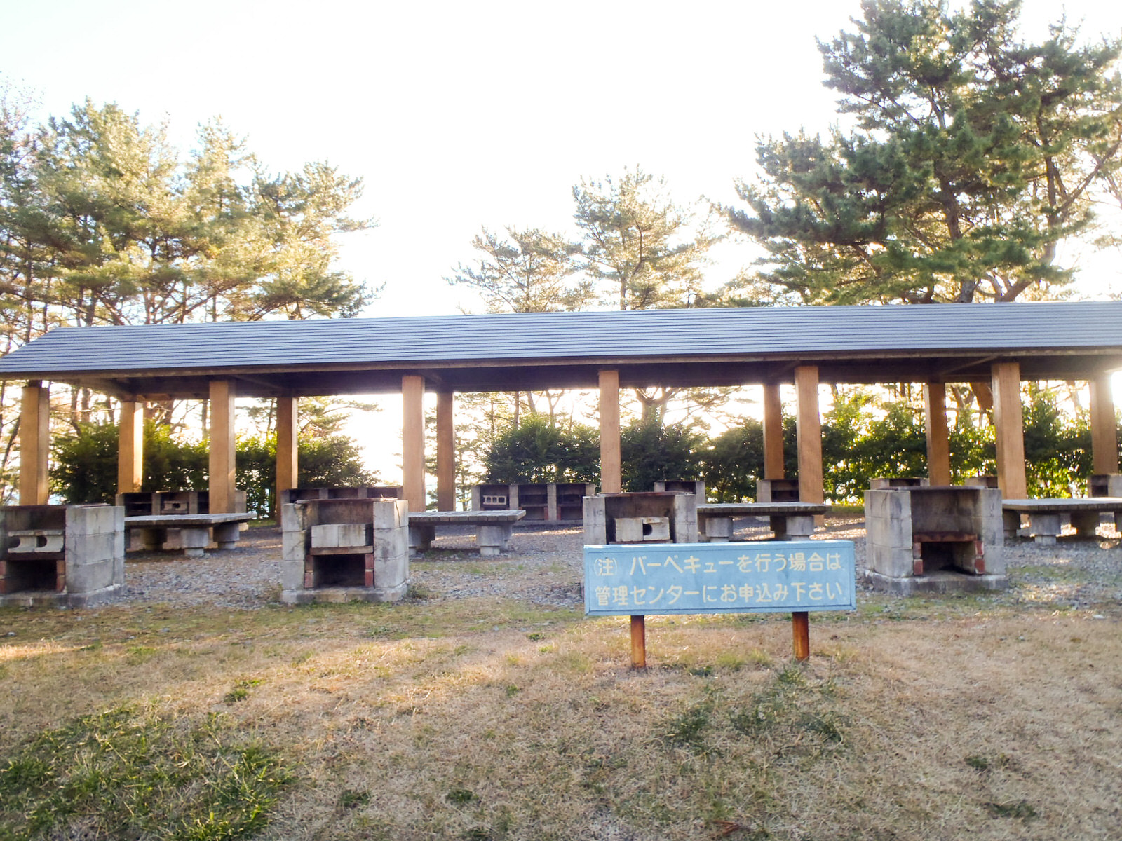 背後には松の木々が立ち並ぶ自然の中で、「バーベキューを行う場合は管理センターにお申込み下さい」と書かれた案内板の後ろに屋根付きの木柱の下にコンクリート製のかまどが整然んだ屋外バーベキュー場の写真