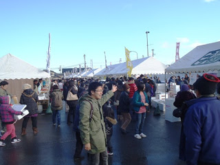 青空の下、白いテントが並ぶ屋外会場で多くの人々が行き交い、出店者や来場者が交流している様子が映された福興市の写真