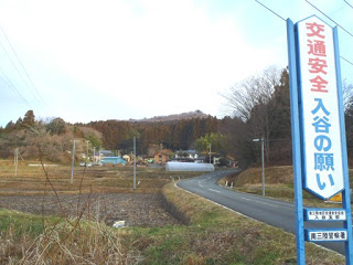 田んぼに囲まれた細い舗装道路のそばに『交通安全 入谷の願い』と書かれた青と白の縦長の看板が立ち、奥に小さな集落と山林が広がっている様子を写した写真