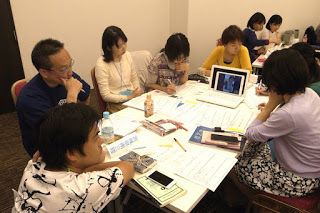 テーブルの上には紙やノート、電子機器が並んだテーブルを囲んで数人が座り、何人かは画像や映像が表示されたノートパソコンの画面を見て話し合っている様子の写真