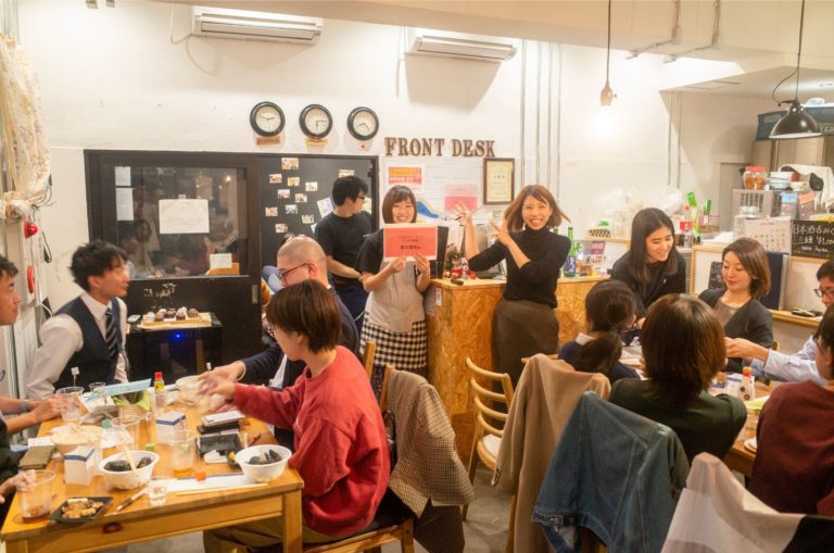 2つのテーブルでたくさんの参加者が食事を楽しむ中、「FRONT DESK」と書かれたカウンターの前に2人の女性が立ち、1人が両手でカウンターの方を示し、もう1人が赤い紙を掲げている様子の写真