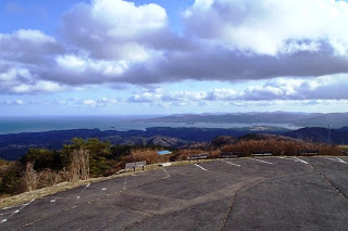 山の上の舗装された駐車場から海と空と広大な山並みを一望できる風景を撮影した写真