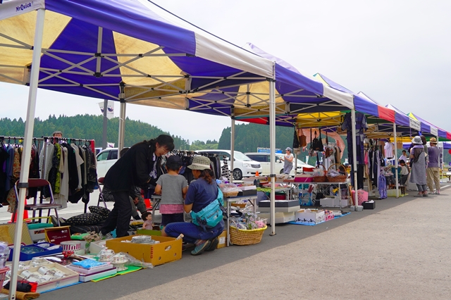 色とりどりのテントが並ぶ青空市場で、人々が衣類や雑貨などの商品を見たり買ったりしてにぎわっている様子を写した写真