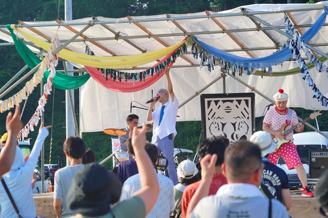 ステージで白いTシャツ姿の男性歌手がマイクを持ち、観客の方に向かって左手を挙げながら熱唱しており、後ろではギターやドラムの演奏者が音楽を奏でている、野外音楽ライブの写真