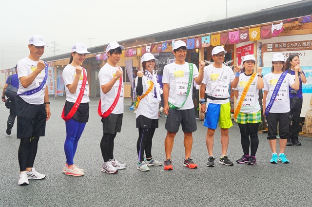 白いキャップを被り、それぞれ紫、赤、黄色、緑、白色のタスキをかけた8名のランナーが横一列に並び、ガッツポーズをして記念撮影をしている写真