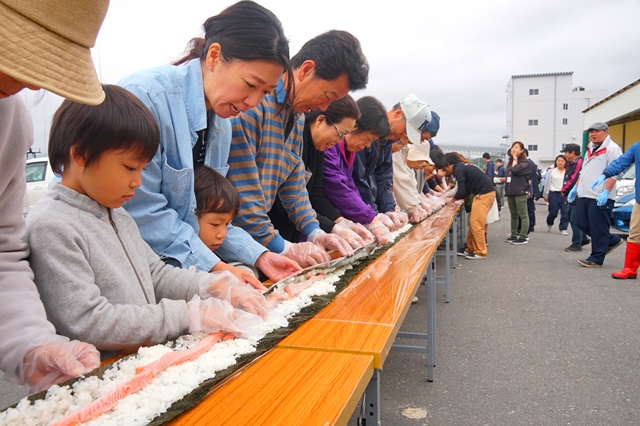 長机を横一列に長く並べ、その上で海苔、酢飯、ギンザケの切り身を巻いたのり巻きづくりが行われている写真