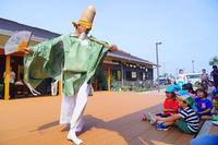 緑の衣装と大きな帽子をかぶった人物が扇子を持って子どもたちの前で舞っている写真