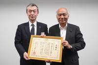 スーツ姿の二人の男性が額縁に入った感謝状を一緒に掲げている写真