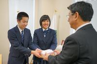 スーツ姿の佐藤町長が右側で背を向け、左側の男子学生と中央の女子学生が笑顔で並び、両手で持った募金箱を町長に手渡している写真