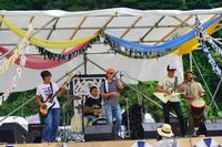 ステージ上で5人の男性がエレキギターやジャンベなどの楽器を演奏し、中央のボーカルがマイクを手に歌っている、野外音楽フェスの演奏中の写真