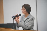 グレーのスーツを着た女性が演台でマイクを持ちながら微笑んで話しており、挨拶をしている様子を写した写真