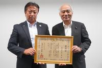 スーツ姿の男性二人が、感謝状の額縁を一緒に持って笑顔でカメラに向かっている写真