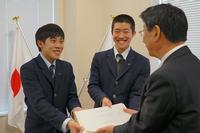 スーツ姿の佐藤町長が右側で背を向け、左側の男子学生と中央の男子学生が笑顔で並び、両手で募金箱を町長に手渡している写真