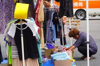 屋外の市場で、ハンガーラックに掛けられた洋服の前に女性がしゃがみ込み、段ボールに入った衣類を丁寧に選んでいる様子を写した写真