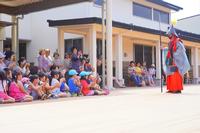 グレーと赤色の衣装の人物が槍を持ち、多くの子どもたちがその前で座って見ている写真