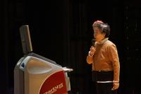 舞台の上でカラオケ機に向かってマイクを持った高齢の女性が歌っている写真