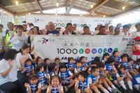 「未来への道 1000キロメートル縦断リレー」と書かれた横断幕の前に、お揃いの衣装を着た子供たち、横断幕の後ろに関係者やランナーたちが並び記念撮影をしている写真