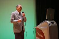 スーツ姿の高齢の男性が舞台の上でカラオケ機に向かって歌っている写真