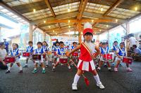 赤い衣装を着た1人の園児が笛を口にくわえメジャーバトンを持ち、その後ろに青い衣装を着て小太鼓を肩から下げた鼓笛隊の園児たちが並んでいる写真