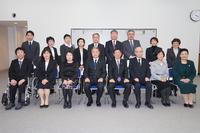 スーツやフォーマルな服装をした大人の男女が二列に分かれて整然と並び、カメラに向かって記念撮影をしている集合写真