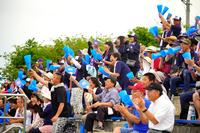 スタンドで青いメガホンを手にした大勢の観客たちが立ち上がり、声援を送りながら高校野球の試合を熱心に応援している写真