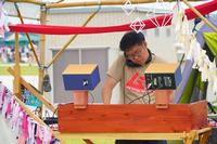 屋外の装飾的なブース内で、Tシャツを着た男性DJがターンテーブルのような機材を操作している、DJパフォーマンスの写