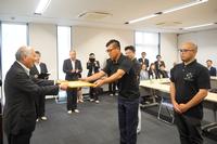 周囲の人々が見守るなか、スーツ姿の男性が感謝状を黒いシャツの男性に手渡している写真