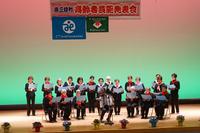 舞台の上の高齢の合唱団が、舞台の中央に座っている男性のアコーディオンの演奏のもと歌を歌っている様子の写真