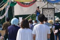 観客が何人か背中を向けてステージを見ており、その先で黒い服の女性がマイクを持って歌っている様子が見える、観客とステージの一体感があるライブ中の写真