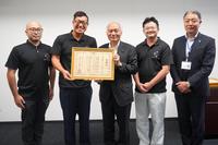 スーツ姿の男性と黒いシャツの男性たちが感謝状を一緒に持ち笑顔で並んでいる写真