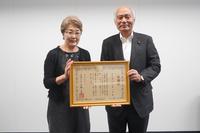 笑顔の男女二人が、部屋の白い壁を背景に、賞状を両手で持ち、カメラに向かっている様子を捉えた写真