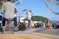 ステージ上から演奏中の様子と観客席、背景の緑の丘が見えるライブ風景の写真