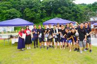 青空の下、イベント会場に設置された紫色のテントの前でグループで集合写真を撮っているイベントスタッフや参加者たちの写真