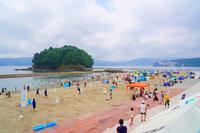 人で賑わう砂浜と、沖合に浮かぶ緑の島、海沿いのイベントテント群が並ぶ夏のビーチイベントの写真