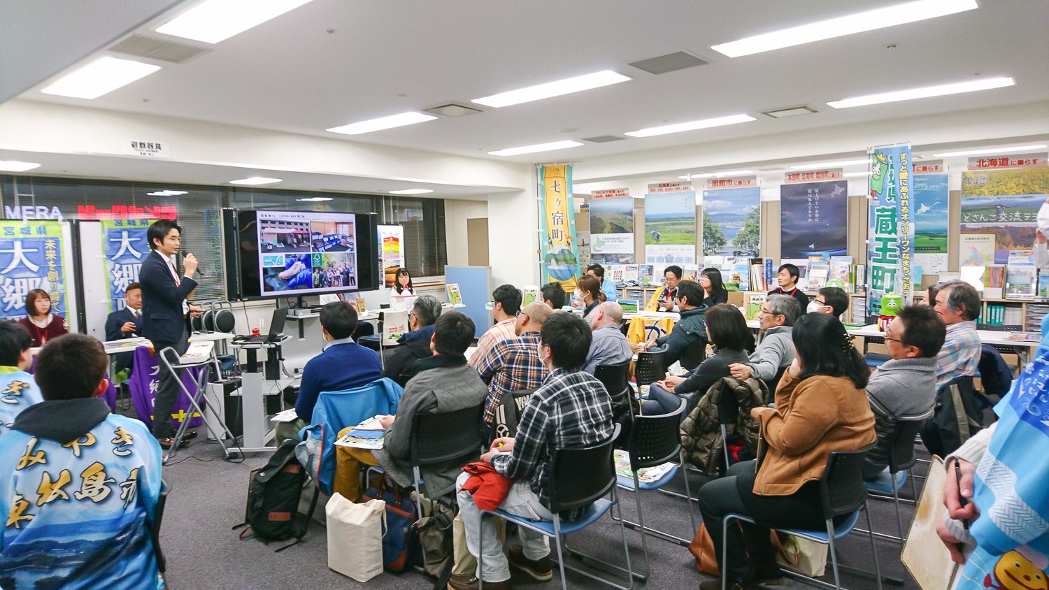 室内のイベント会場で、多くの人々が椅子に座り、スーツ姿の男性のプレゼンテーションを真剣に聞いている様子の写真