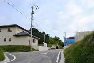 ゆるやかな坂道の左右に住宅が建ち並び、電柱や工事車両が見える住宅街の建設途中の写真