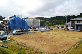 手前に家の基礎部分の跡がある更地が広がり、奥では数棟の住宅が建設中で工事車両が停まっている造成地の写真