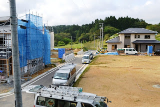 左側の建設中の建物には足場と青い防塵シートがかかり、右奥には完成した住宅と並ぶ車が見える造成地の写真