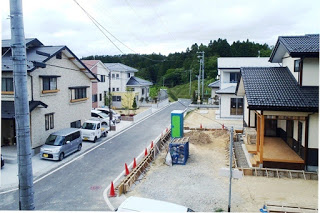 舗装された道の両側に住宅が建ち並び、前方には山林が広がる静かな住宅街の写真