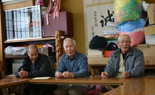 地元のわら細工名人である三人の高齢男性が机の前に座っている写真