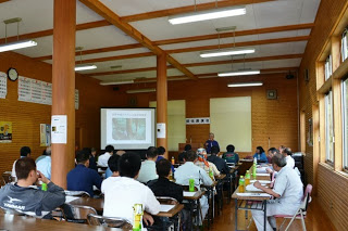 木造の会議室で前方のスクリーンに映像が映し出され、多くの参加者がに席に座って真剣に講演会の話を聞いている様子の写真