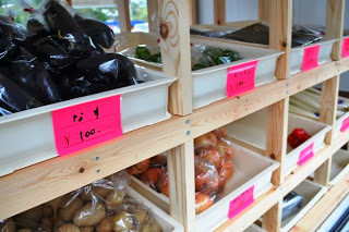 木の棚に並べられた白いトレーの中にナスやピーマン、玉ねぎ、ジャガイモなどの袋詰め野菜が手書きの値札と共に並べられている写真