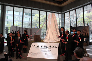 屋内の広いガラス張りのスペースに展示された、「Moai 未来に生きる」と書かれた台座に乗ったモアイ像にかぶせた白い布からのびた赤い紐を、周りに立ったスーツ姿の男性や制服姿の高校生が持っている除幕式の記念写真